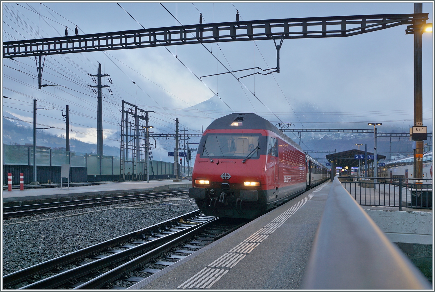 Die SBB Re 460 004 (UIC 91 85 4 460 004-5 CH-SBB) wartet in Brig mit einem IR 90 auf die Abfahrt nach Genève Aéroport. 

3. Jan. 2024